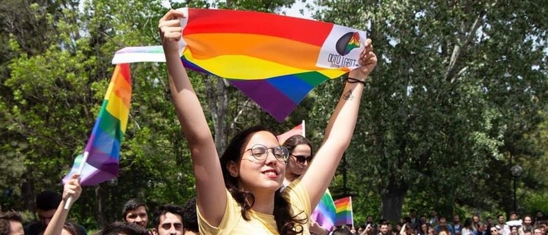 A student wearing thin rimmed glasses holds up a rainbow flag. They are surrounded by people holding similar flags. 