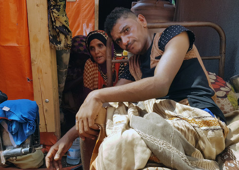 A young man looks intently at the camera. He is sitting on a small metal bed, his legs draped in light coloured woven blankets and fabric. A woman sits on the floor behind him.