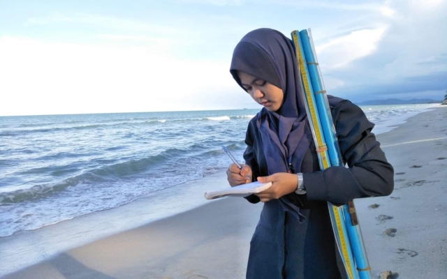 Khairiyah Rahmanyah stands on the beach and is writing into a notebook.