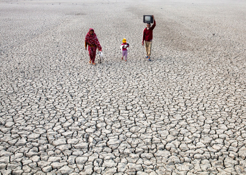 A family walks across a dried river bed. The ground is made up of deeply cracked, white soil that continues past the horizon. The family includes a woman wearing pink fabric that drapes over her head, a young child carrying a small white goat and a man wearing a red jumper and carrying a television set.