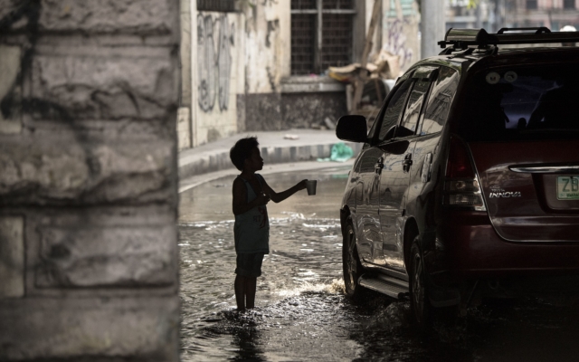 a young boy stands, feet covered in water, holding a cup out to a car as it drives past