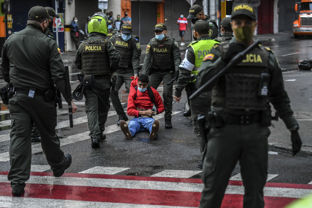 A group of police pull a young man in a red jumper and a surgical mask is pulled across the ground by the hood of his jumper. 