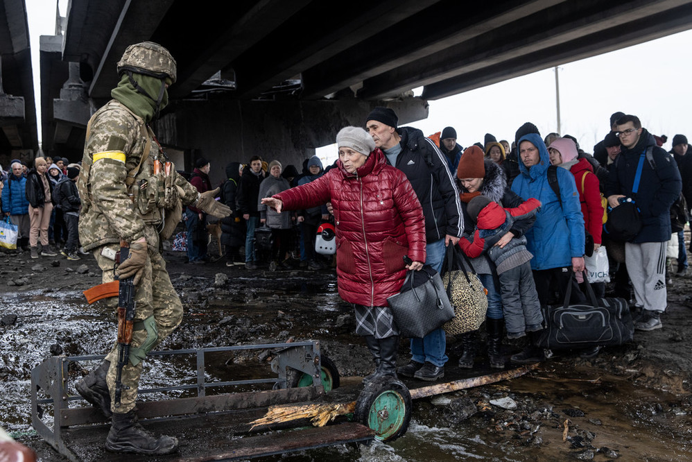 A group of people fleeing their homes due to the conflict in Ukraine assemble near a bridge. A solider in uniform helps an older person cross walk across muddy ground.