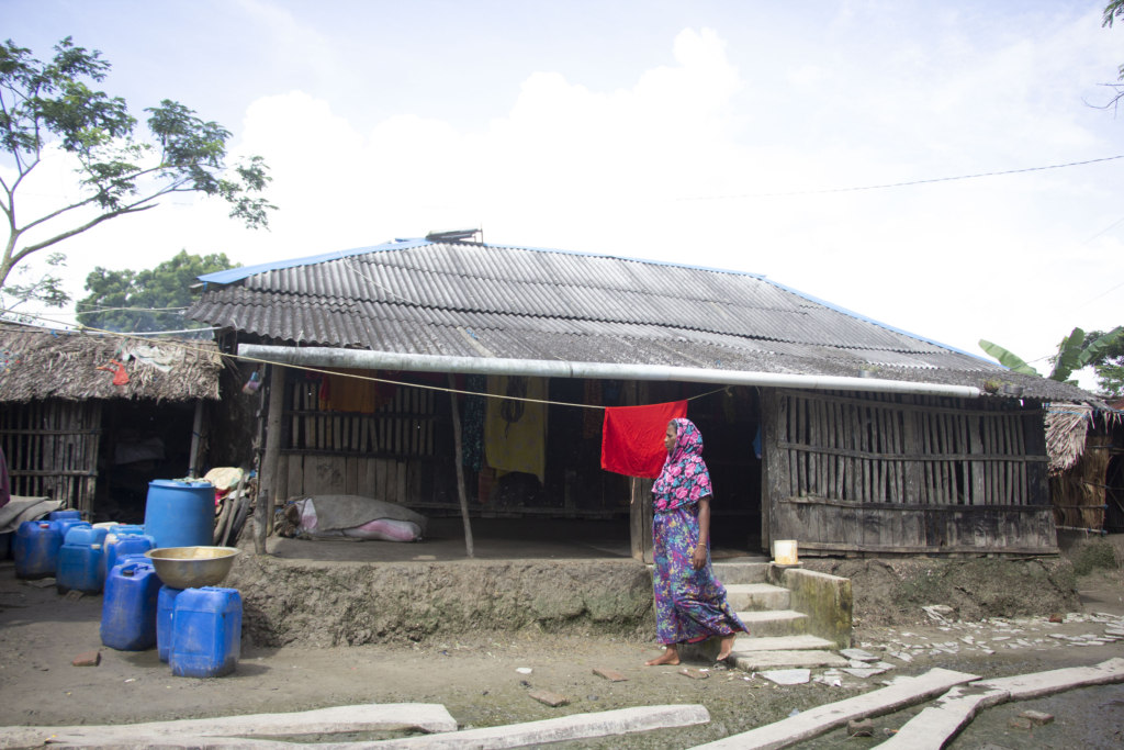 a woman wearing a pink and purple dress walks outside a building. The building has a rough tin roof and walls built by wooden slats that have holes in them. You can see blue containers in on the left which are likely filled with water.