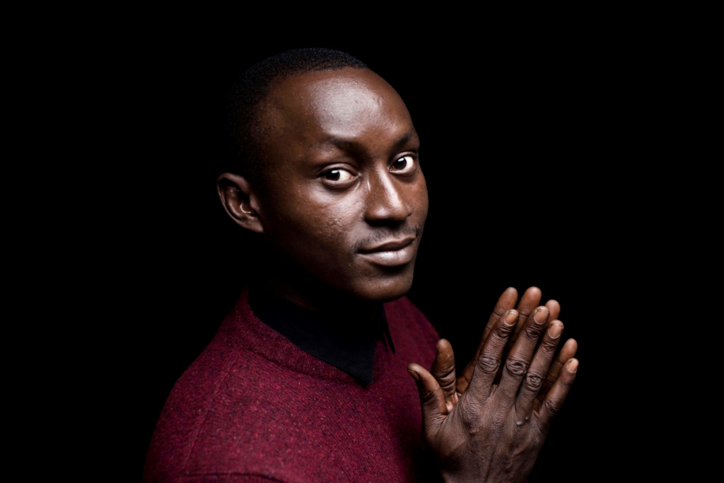 a portrait of Moses Akatugba. He wearing a red shirt and is holding his hands together. 