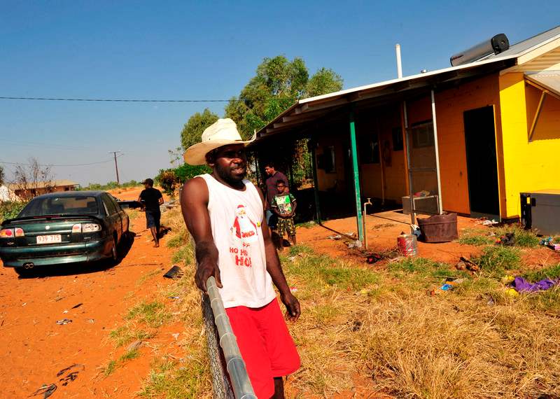 An image of Rosco who is leaning on a metal fence in front of a house. He is wearing red shorts, a white t shirt and a white woven wide brimmed hat that shades his face.