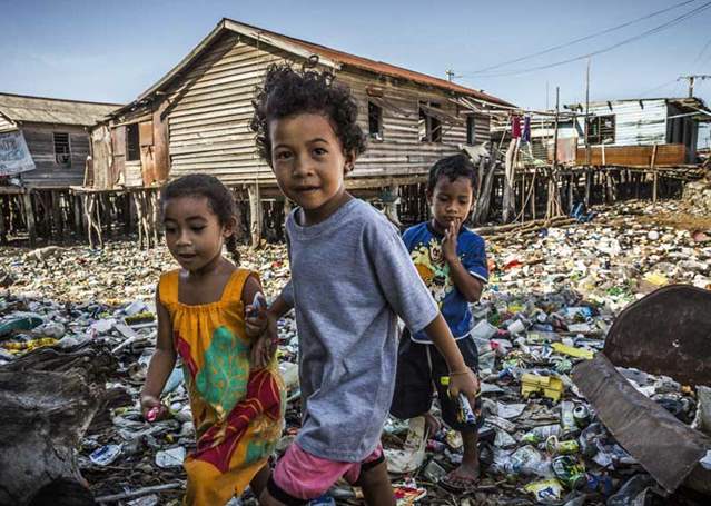 three young children walk across a rubbish dump. Behind them is a small house held up by stilts