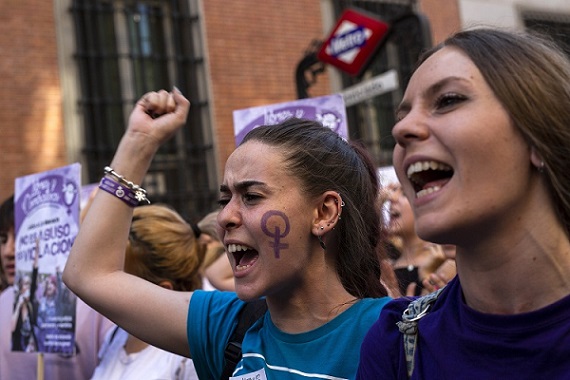 two women are in focus in the image, both look as though they are speaking or chanting. The one on the left has a Venus symbol painted in purpose on her cheek and is holding up a fist.